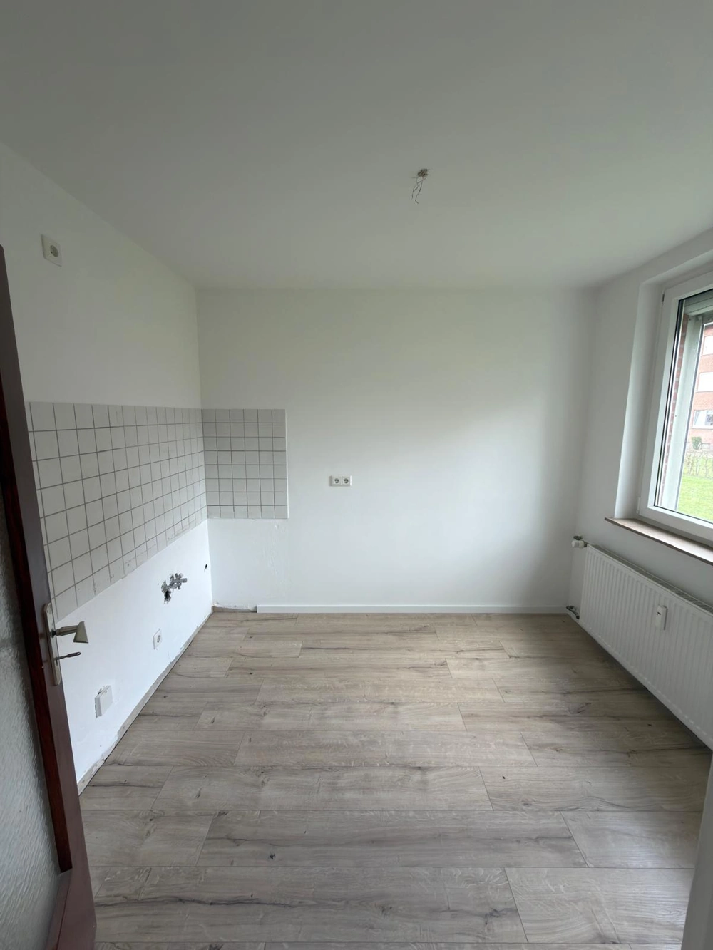 Leerer Raum mit neu verlegtem Laminatboden, weißer Fliesenrückwand, Wasseranschlüssen und Heizkörpern – Küchenrenovierung