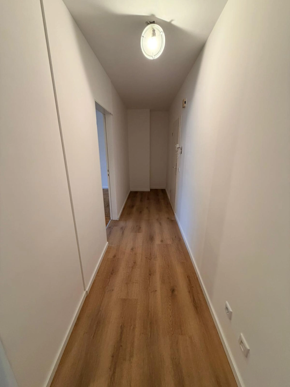Schmaler Flur nach Laminat verlegen mit weiß gestrichenen Wänden, Holzoptikboden und Deckenleuchte