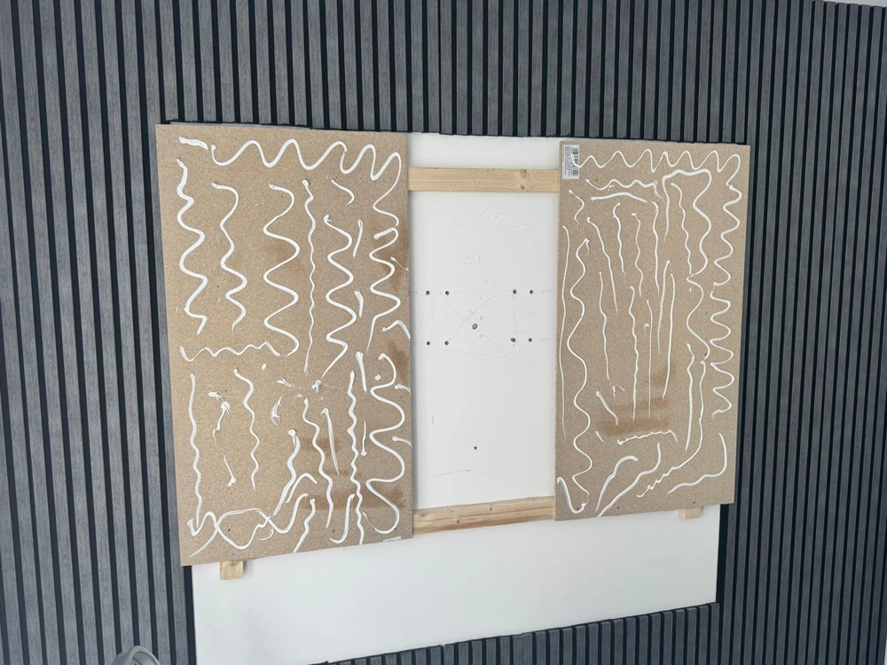 Zwei Spanplatten mit aufgetragenem Klebstoffmuster auf Holzunterkonstruktion vor dunkler Lamellenwand, Trockenbau
