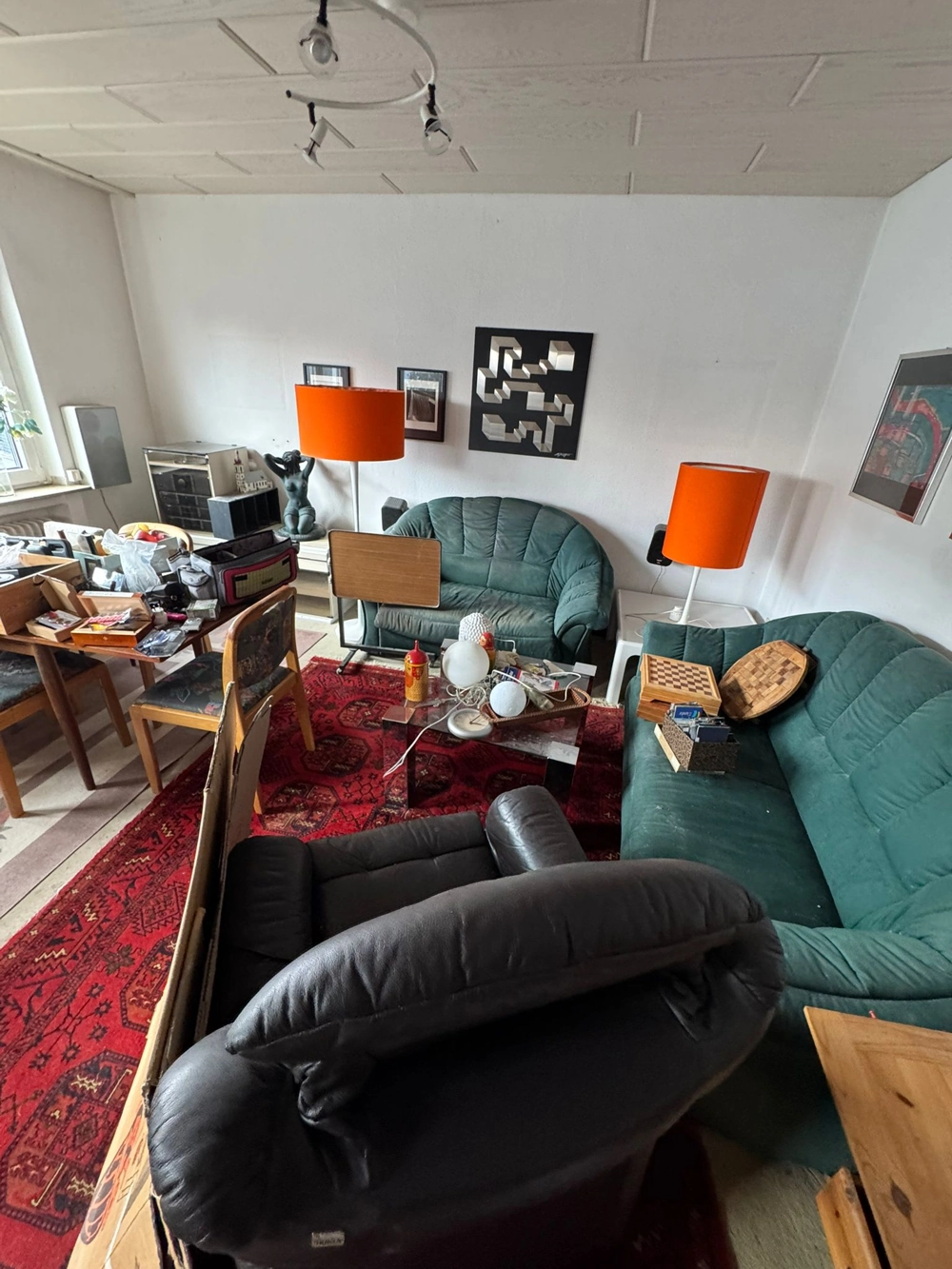 Vollgestelltes Wohnzimmer mit grünen Sofas, rotem Teppich, orangen Stehlampen und Kisten vor Entrümpelung