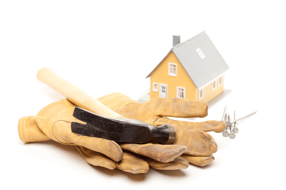 Hammer und Lederhandschuhe neben Nägeln und einem Hausmodell als Werkzeug für Trockenbau