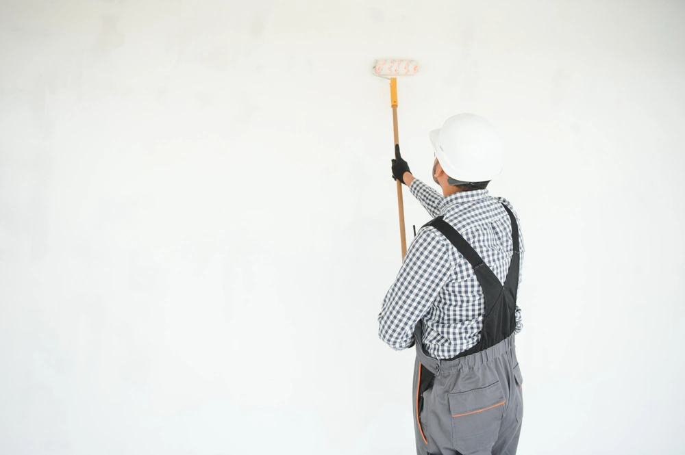 Handwerker mit Schutzhelm und Handschuhen streicht weiße Innenwand mit Farbrolle auf Teleskopstiel bei Malerarbeiten