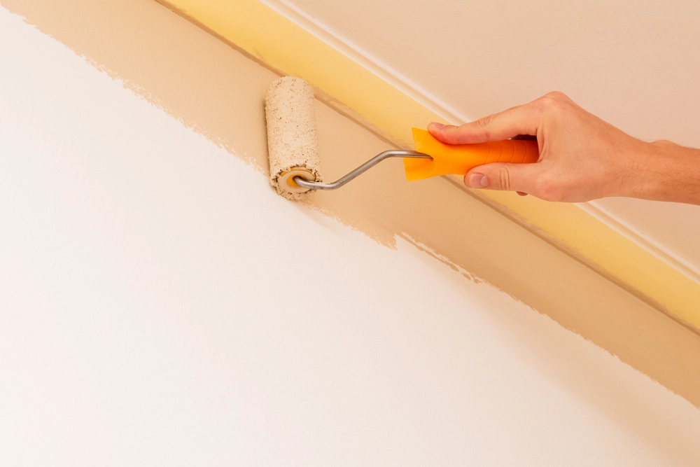 Hand mit Farbroller streicht Wandkante hellbeige unter Deckenleiste, Malerarbeiten